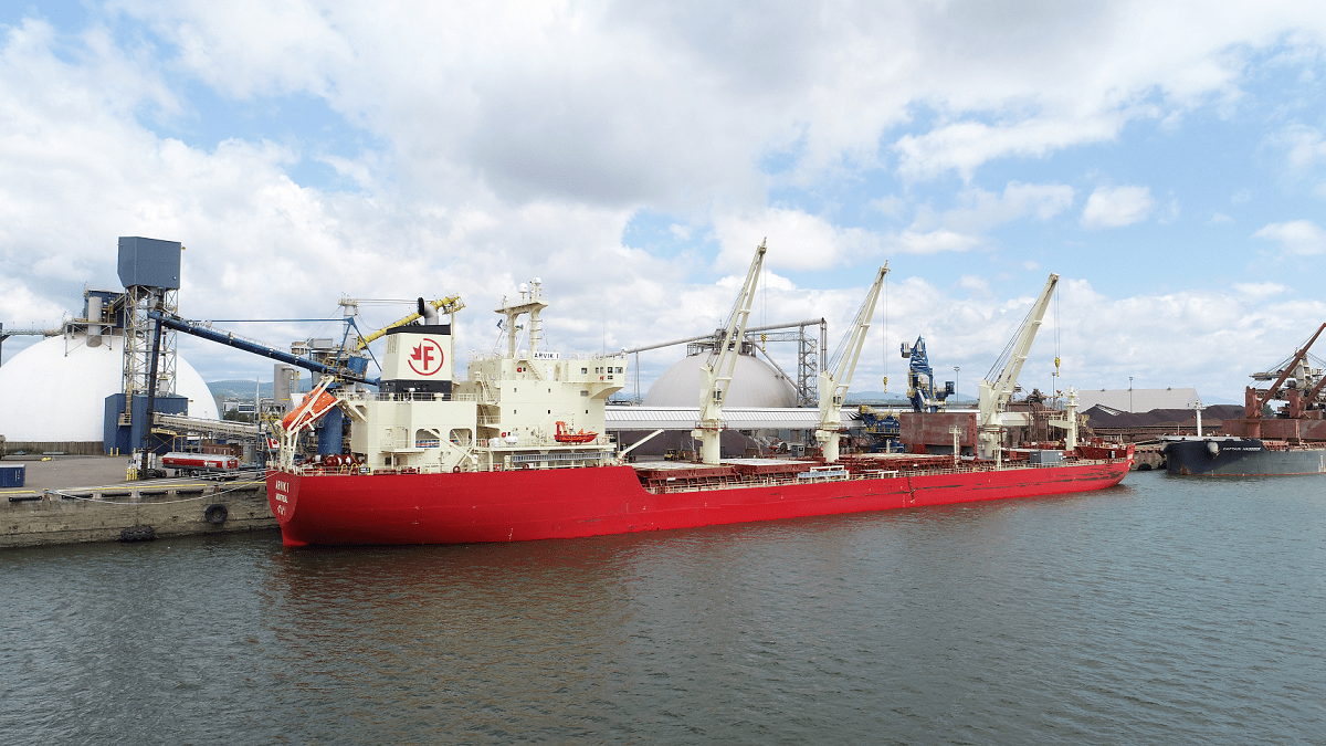 A bright red bulk carrier ship, the MV Arvik I, is docked at an industrial port facility under a partly cloudy sky. Large white storage domes and multiple conveyor systems are visible in the background, along with cranes used for loading and unloading cargo.